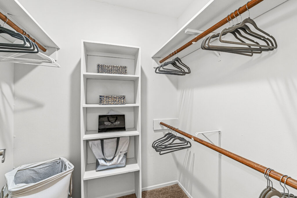 Empty walk-in closet at Enclave at Eagle Creek featuring white shelves, wicker baskets, black hangers on wooden rods, and a laundry hamper on the left side.