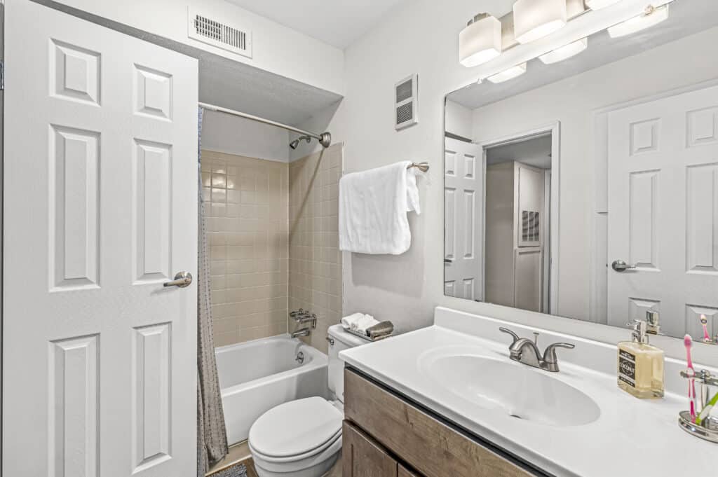 A small bathroom at Enclave at Eagle Creek with a bathtub and shower, toilet, vanity with sink, mirror, wall-mounted light, white and beige tones, a towel on the rack, and toiletries neatly arranged on the counter.