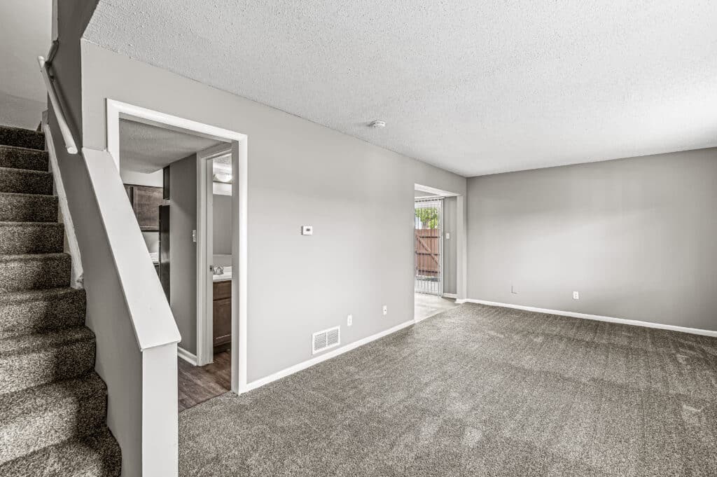 Unfurnished living room in Oakbrook Village - Indianapolis Rentals with carpeted floor, neutral walls, staircase on the left, doorway to bathroom, and sliding glass door leading outside.