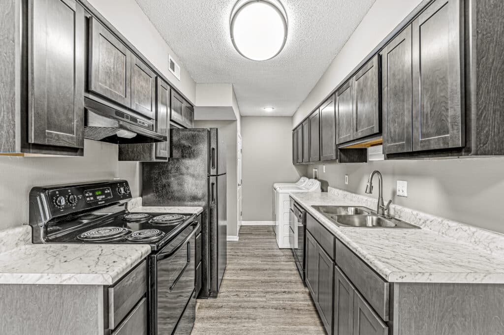 A modern galley kitchen with dark cabinets, black appliances, white marble countertops, and a washer and dryer at the far end—discover stylish living at Oakbrook Village - Indianapolis Rentals.