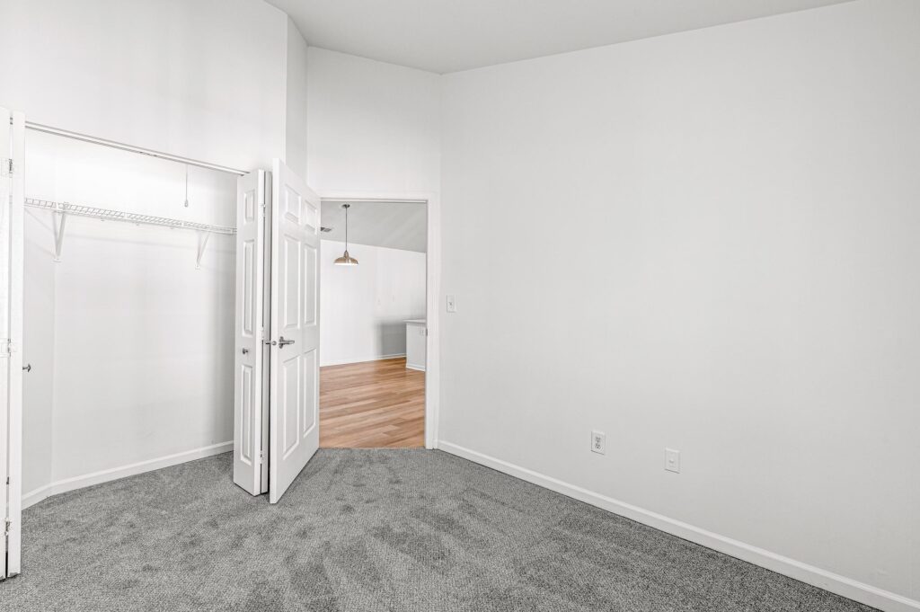 Empty room with white walls, grey carpet, an open closet with shelving, double doors, and a view into a room with wood flooring and a hanging light fixture.