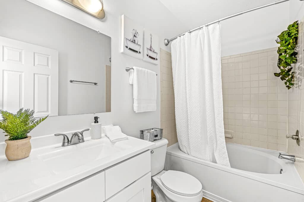 Bright white bathroom at Buffalo Creek Apartments features a sink, mirror, toilet, and bathtub with shower curtain; beige tiled walls, stylish towel, wall art, and lush potted plants complete the inviting space.