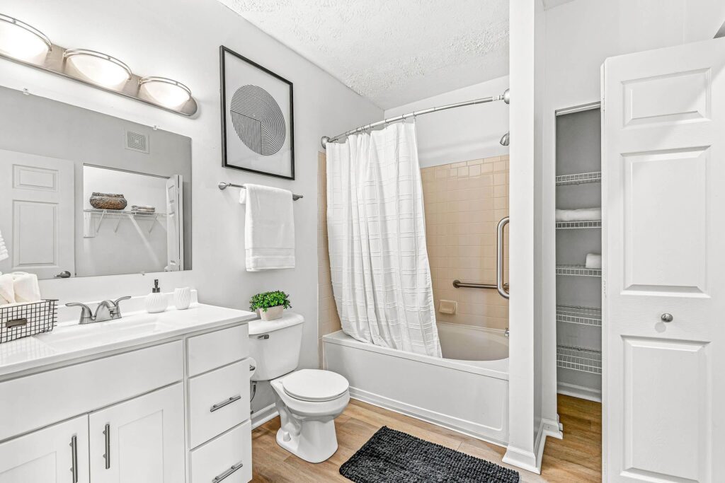 A clean, modern bathroom with white cabinets, a sink, toilet, bathtub with a shower curtain, wall art, and open shelving with towels and storage.