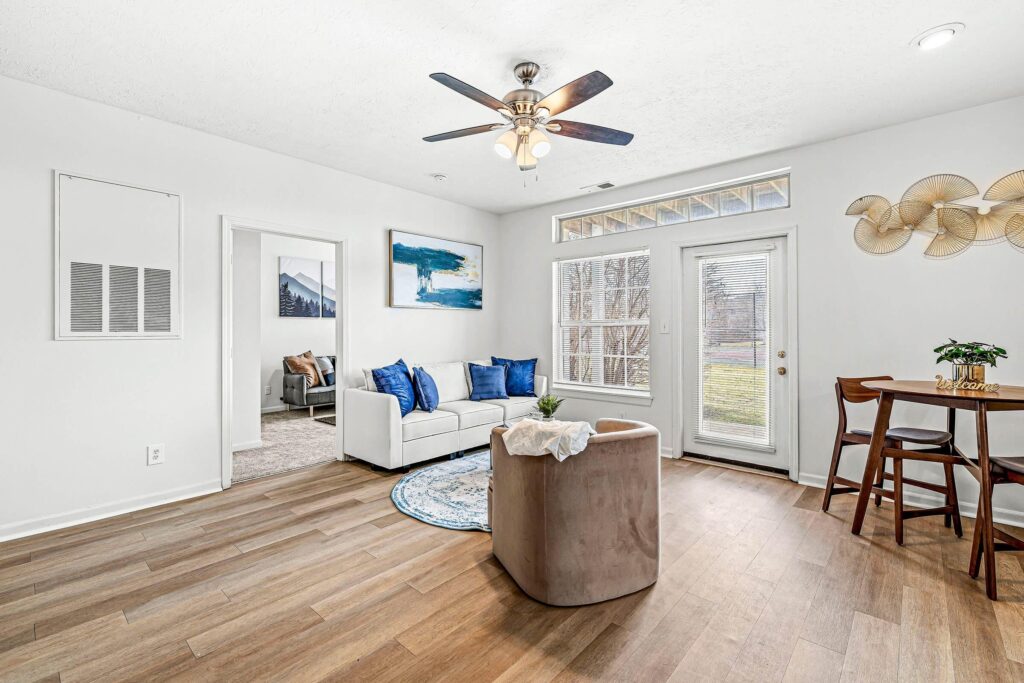 Modern living room with wood flooring, a white sectional sofa, a round chair, a small dining table, wall art, ceiling fan, and large windows with a view of the outdoors.