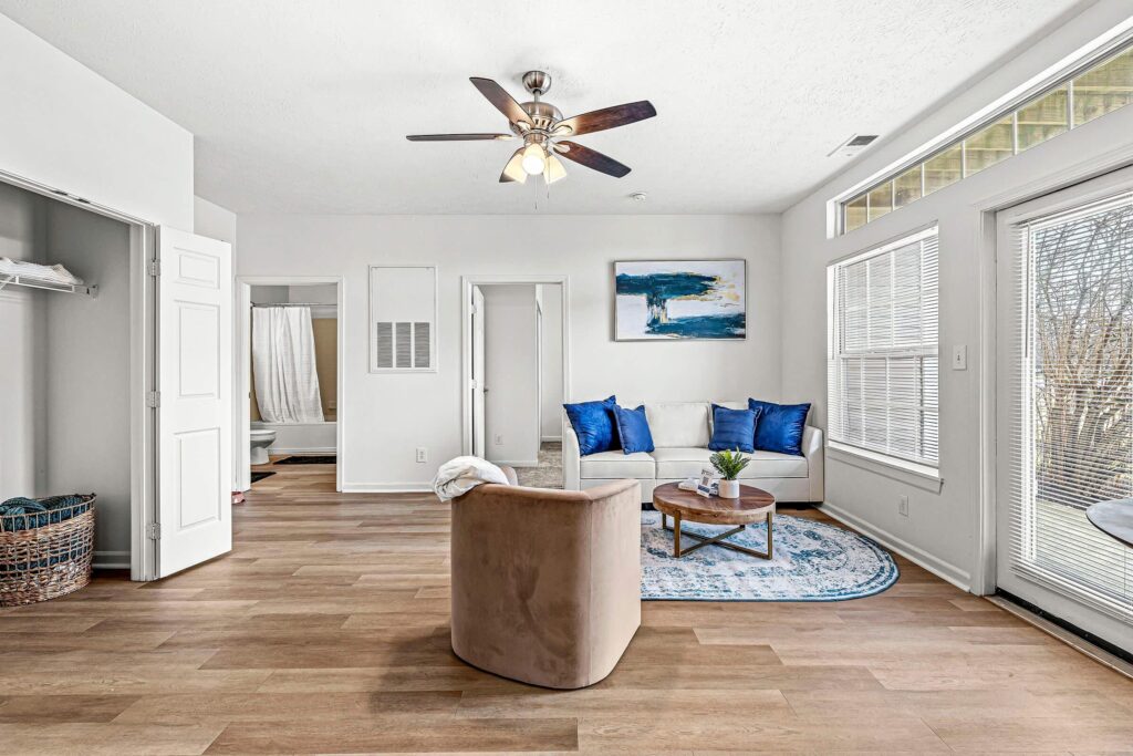 Bright living room with wood flooring, a ceiling fan, a white couch with blue pillows, a round coffee table, and large windows letting in natural light. A bathroom and closet are visible.