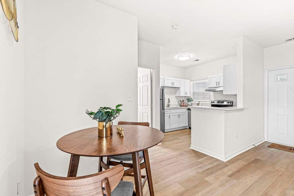 A small dining area with a round wooden table and two chairs is adjacent to a modern white kitchen with stainless steel appliances and light wood floors.