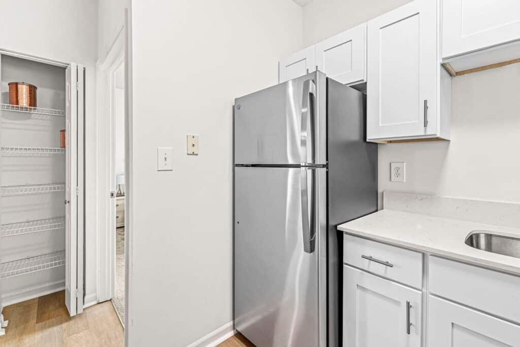 Modern kitchen with stainless steel refrigerator, white cabinets, marble countertop, and an open pantry with wire shelving.