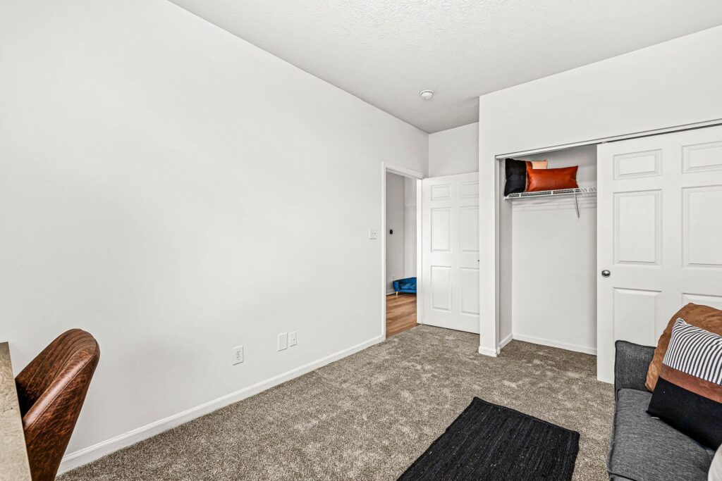 A simple, carpeted bedroom with white walls, a closet with sliding doors, an office chair, a small sofa, and pillows. Door is open to a hallway with wood flooring.