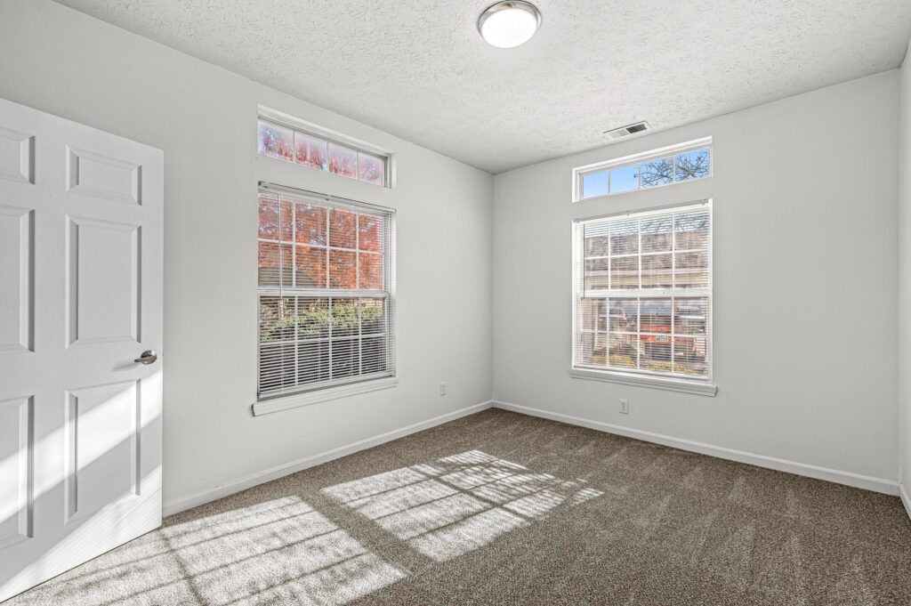 Empty room with white walls, carpeted floor, two large windows, and a closed white door; sunlight casts shadows on the floor.