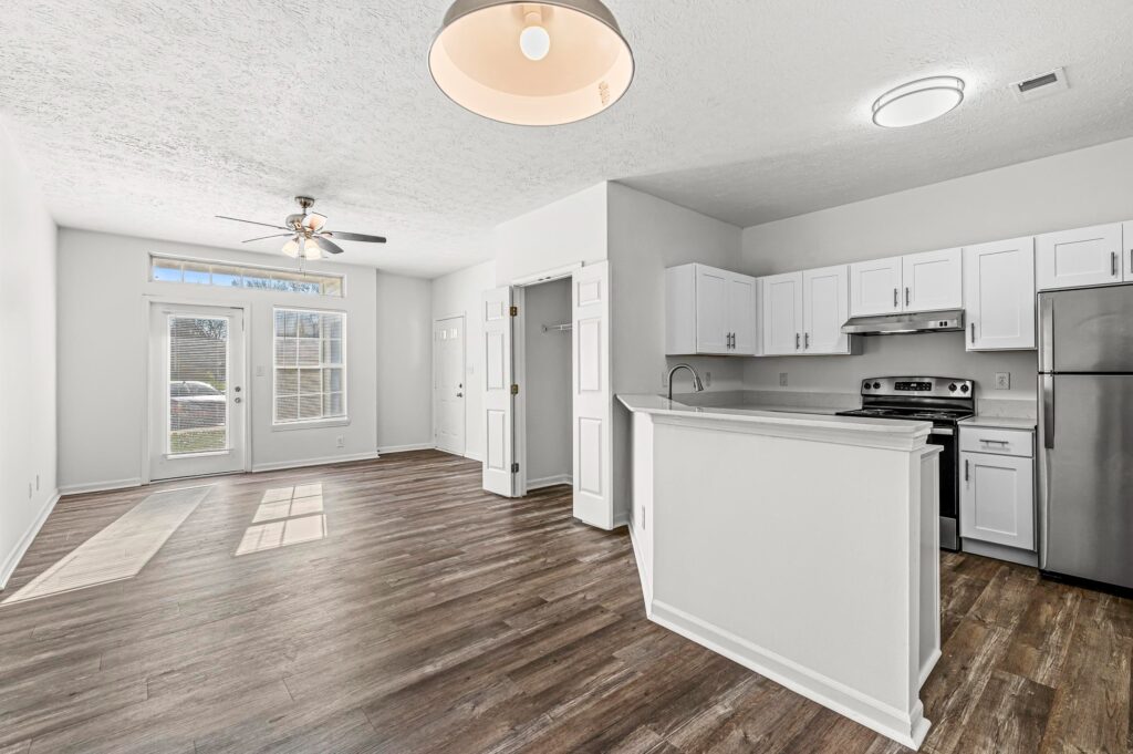 Open-plan living area with wood flooring, white walls, a modern kitchen with stainless steel appliances, ceiling fan, and double glass doors leading outside.