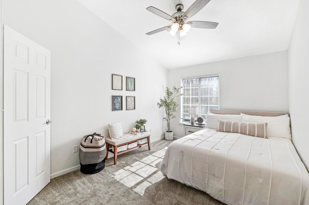 Bright bedroom with a neatly made bed, small bench with pillows, potted plant, wall art, and a ceiling fan; sunlight streams through a window with blinds.