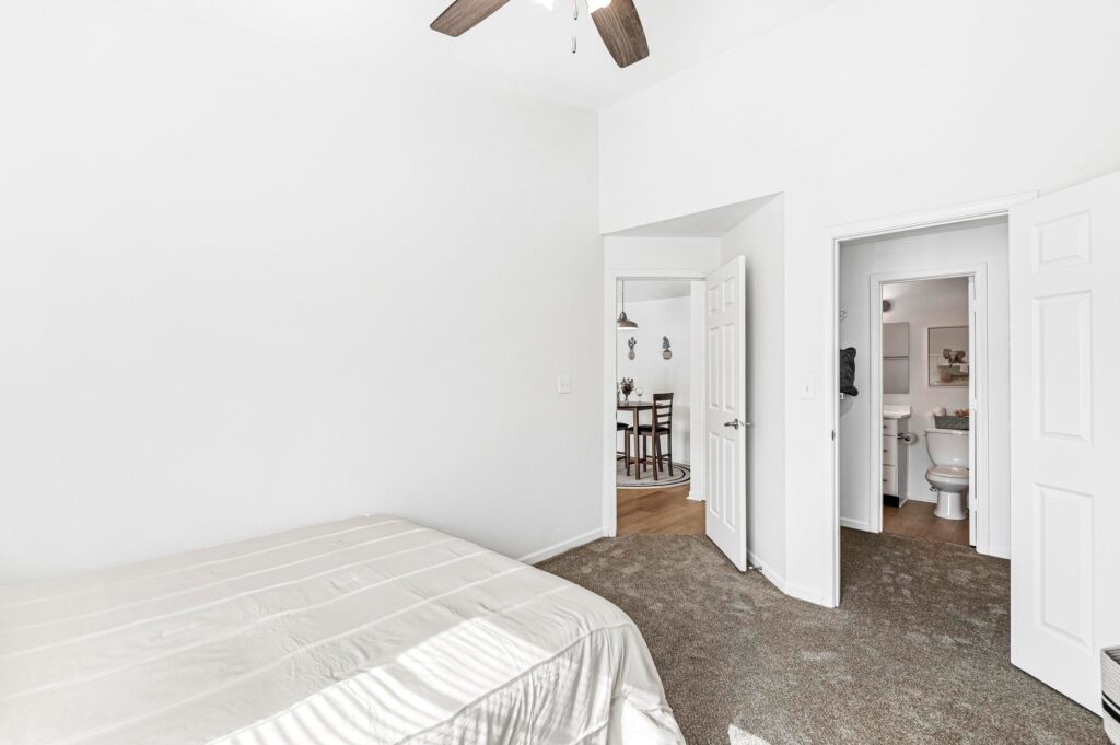 A simple bedroom with a bed, beige carpet, and white walls; doors open to a bathroom and a dining area in the background.