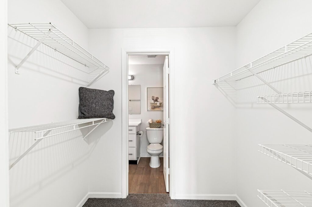 A white walk-in closet with wire shelves on both sides, a single gray pillow on the left shelf, and an open door leading to a bathroom with a toilet and vanity.