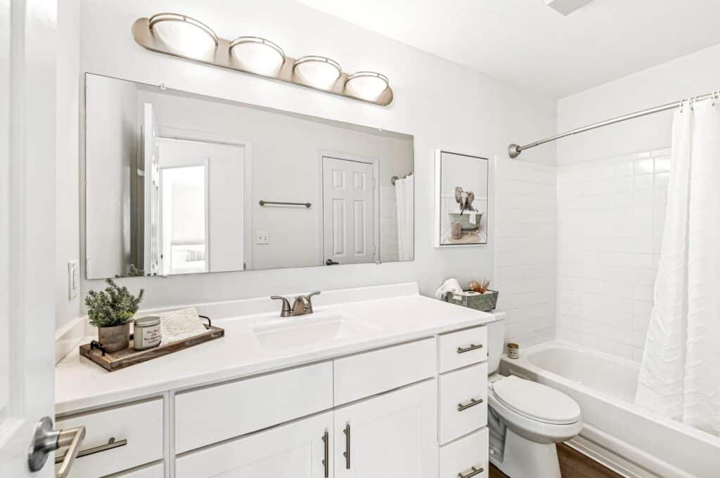 Bright, modern bathroom with white cabinets, a large mirror, bathtub with white tile, and simple decor on the countertop.