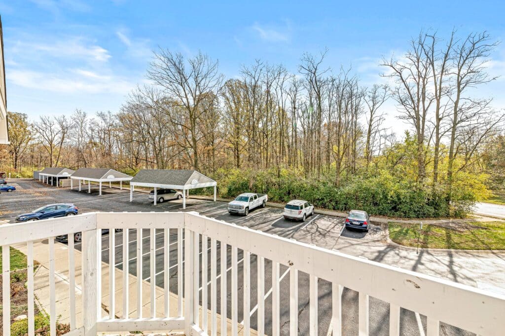 View from a balcony overlooking a parking lot with several cars, carports, and trees in the background on a clear day.