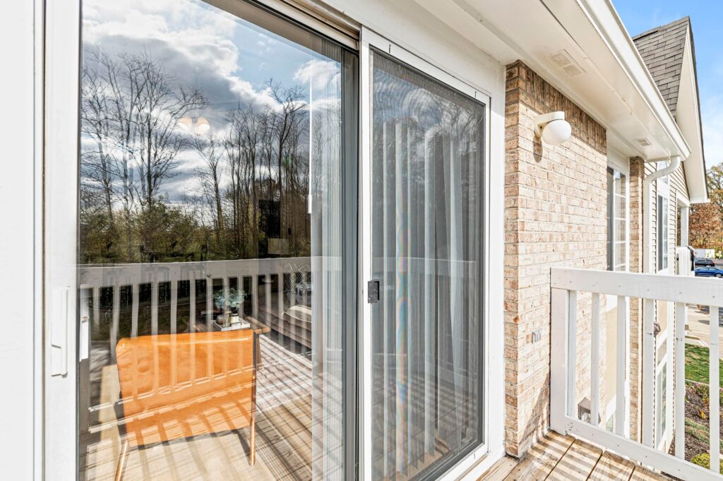 A glass sliding door opens to a small balcony with a wooden chair, white railing, and brick exterior walls. Trees and clouds are reflected in the glass.