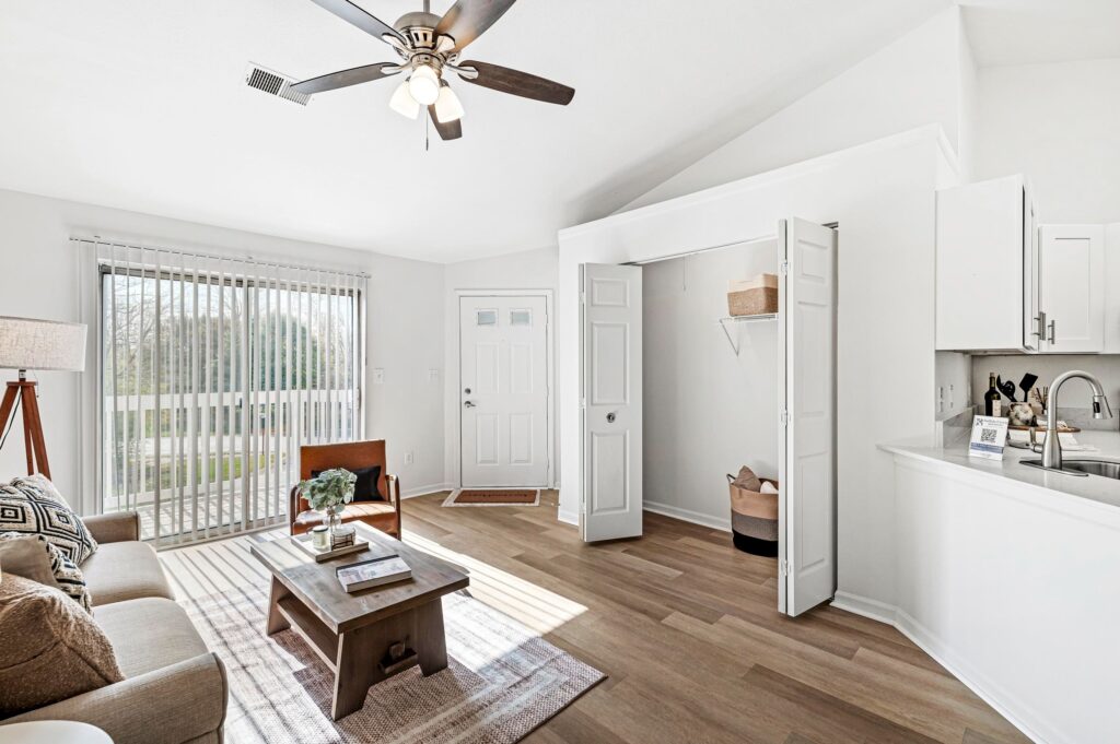Bright living room with a ceiling fan, sofa, coffee table, and open closet near the front door; sliding glass doors let in natural light.