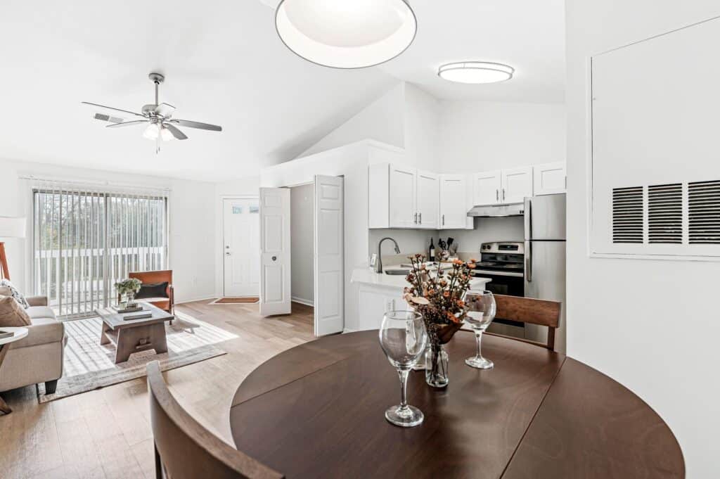 Modern open-concept apartment with a dining table in the foreground, kitchen with stainless steel appliances, and a living area with a sofa near sliding glass doors.