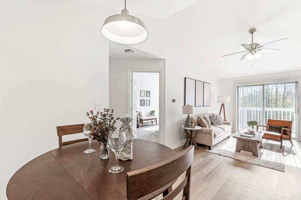 A modern, bright living and dining area with a round wooden table, set for two, and a cozy living room with a sofa, chairs, and large windows letting in natural light.