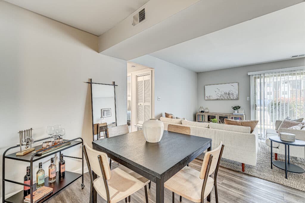 A modern open-plan living and dining area at The Oasis Apartments features a black dining table, light wood chairs, bar cart, sofa, and sliding glass doors leading outside.