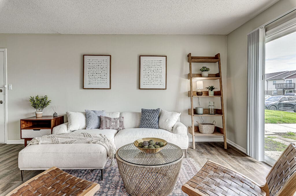A modern living room at Stonehedge Apartments features a white sofa, ottoman, wooden ladder shelf, coffee table, and a large window overlooking a grassy outdoor area.