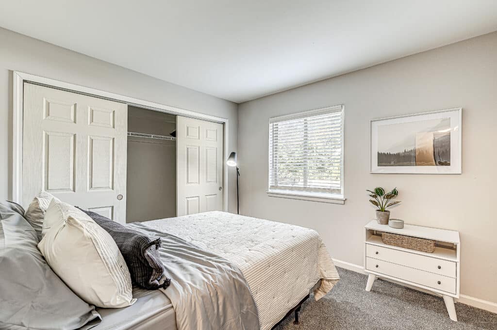 A neatly made bed in a bright bedroom at The Oasis Apartments, featuring gray walls, an open closet, a window with blinds, a small dresser, and a framed picture on the wall.