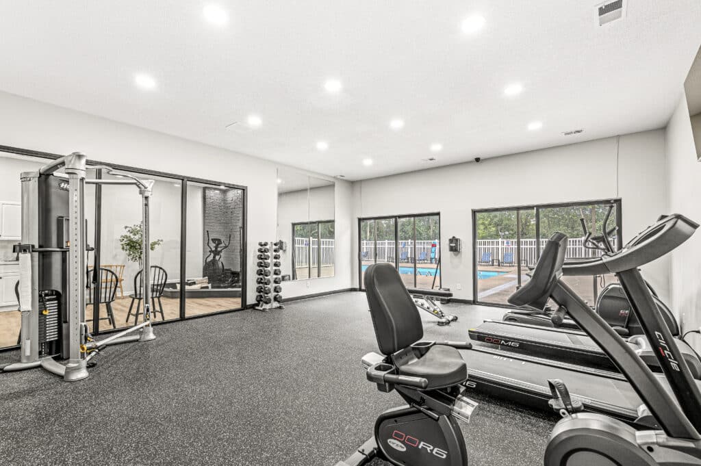 Modern fitness center at The Oasis Apartments with exercise bikes, treadmills, weight machine, free weights, and large windows overlooking an outdoor pool area.