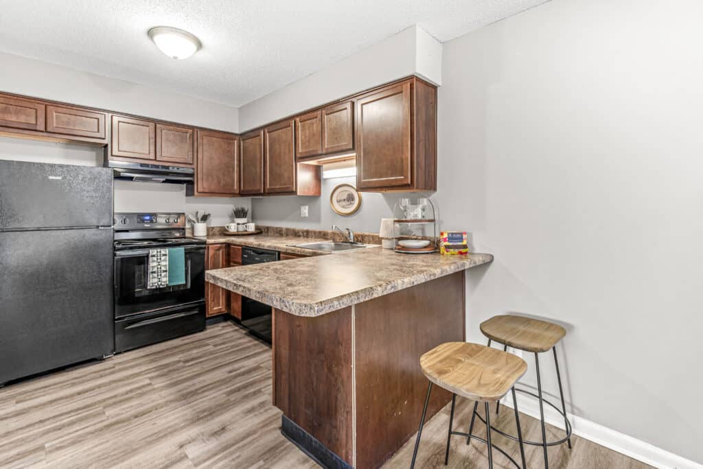 Modern kitchen at Oakbrook Village Apartments featuring dark wood cabinets, black appliances, and a breakfast bar with two wooden stools, set on a light wood floor and neutral walls.