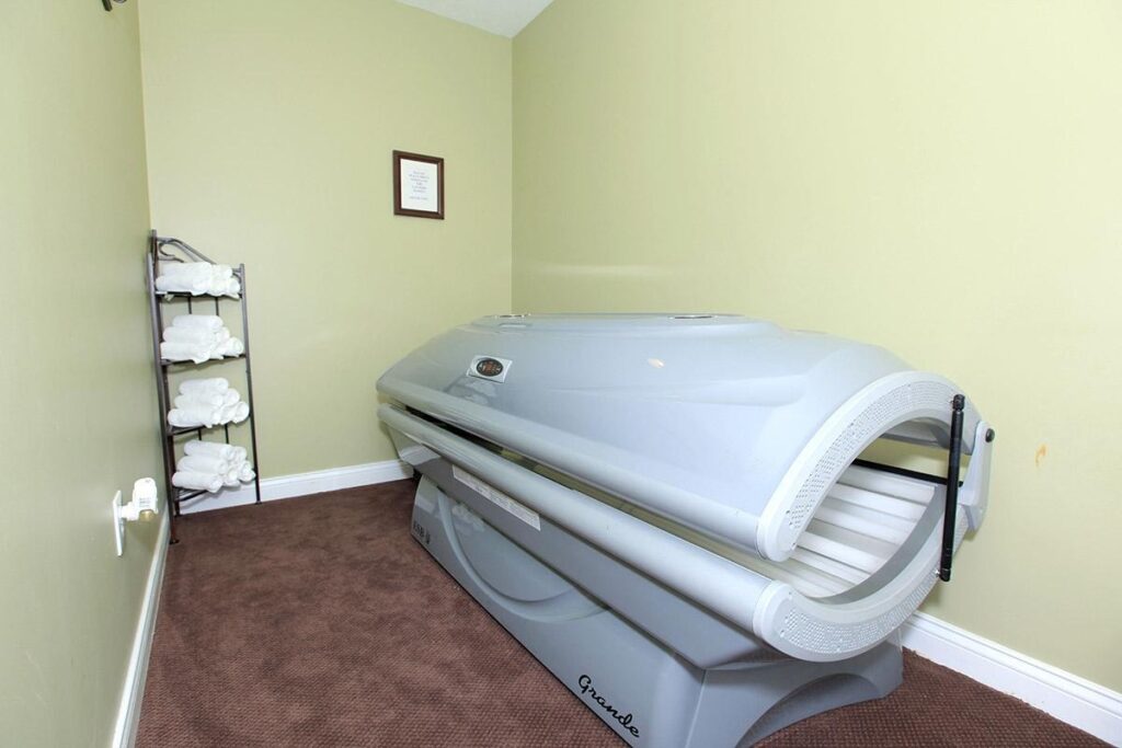 A tanning bed is positioned in a small room with light green walls, brown carpet, and a shelf holding neatly folded white towels.