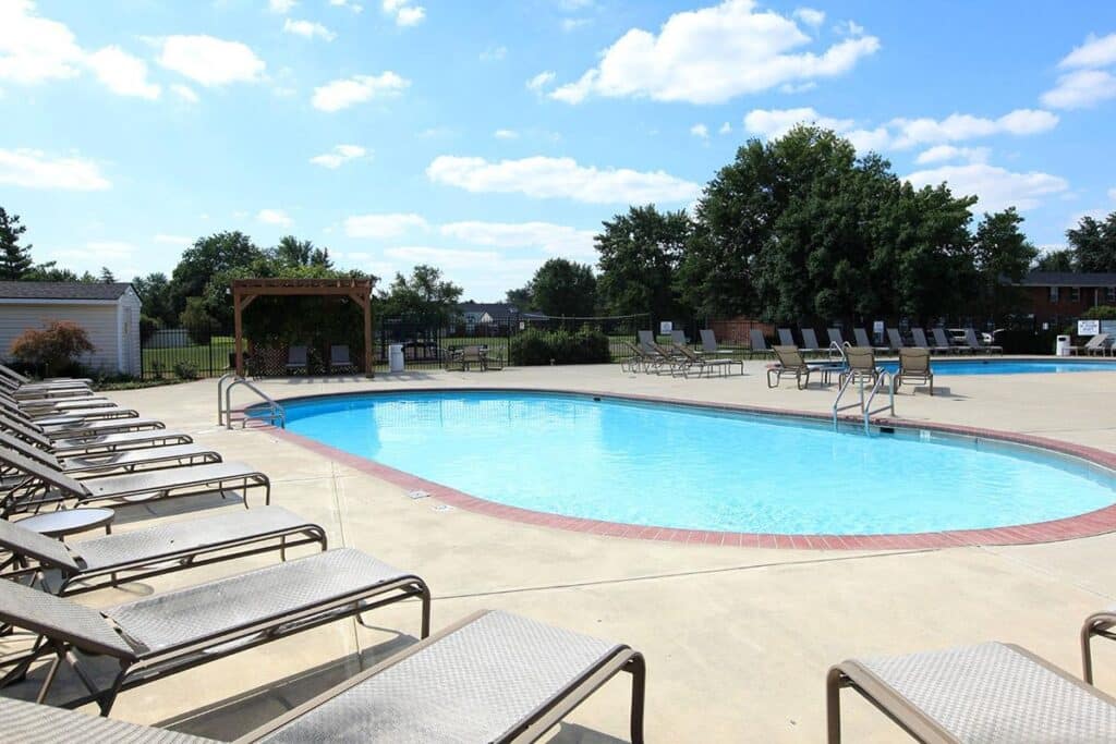 Outdoor swimming pool surrounded by empty lounge chairs on a sunny day, with trees and a few buildings in the background.