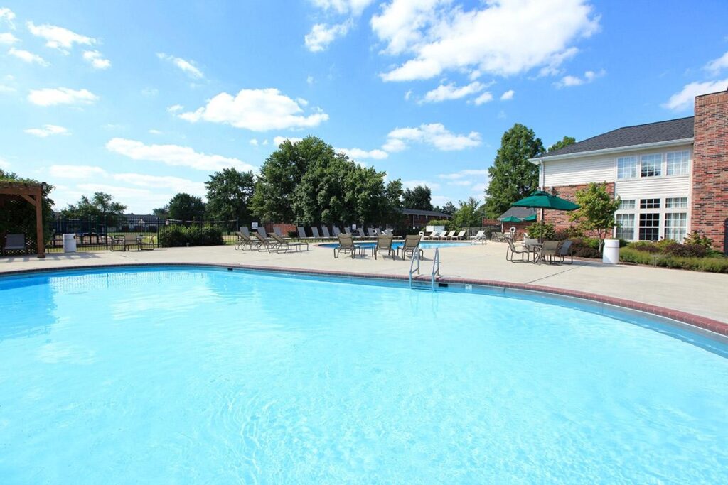 Outdoor swimming pool with clear blue water, surrounded by lounge chairs, umbrellas, trees, and a brick building under a sunny sky.