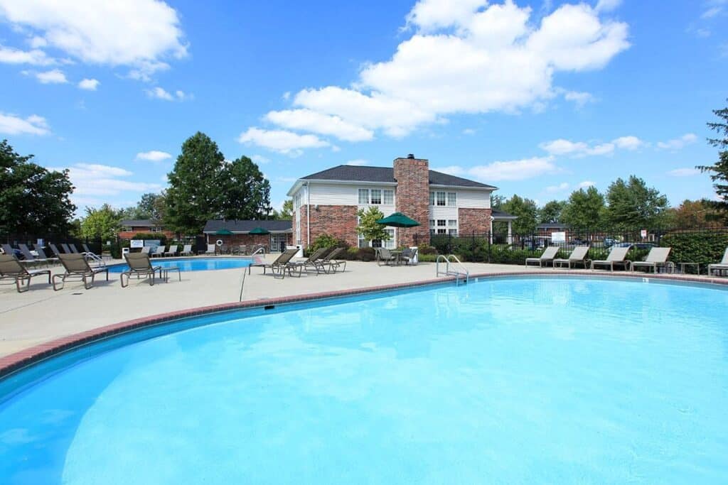 Outdoor swimming pool with clear blue water, surrounded by lounge chairs and umbrellas, with a brick building and trees in the background under a partly cloudy sky.