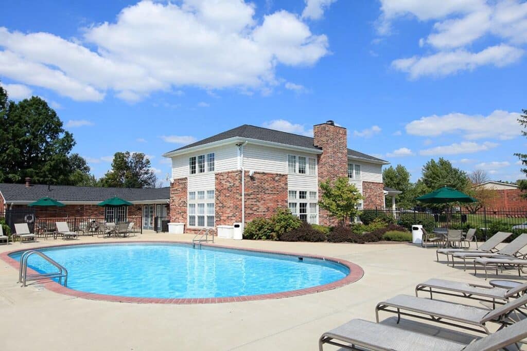 Outdoor swimming pool surrounded by lounge chairs, umbrellas, and a two-story brick building under a partly cloudy sky.