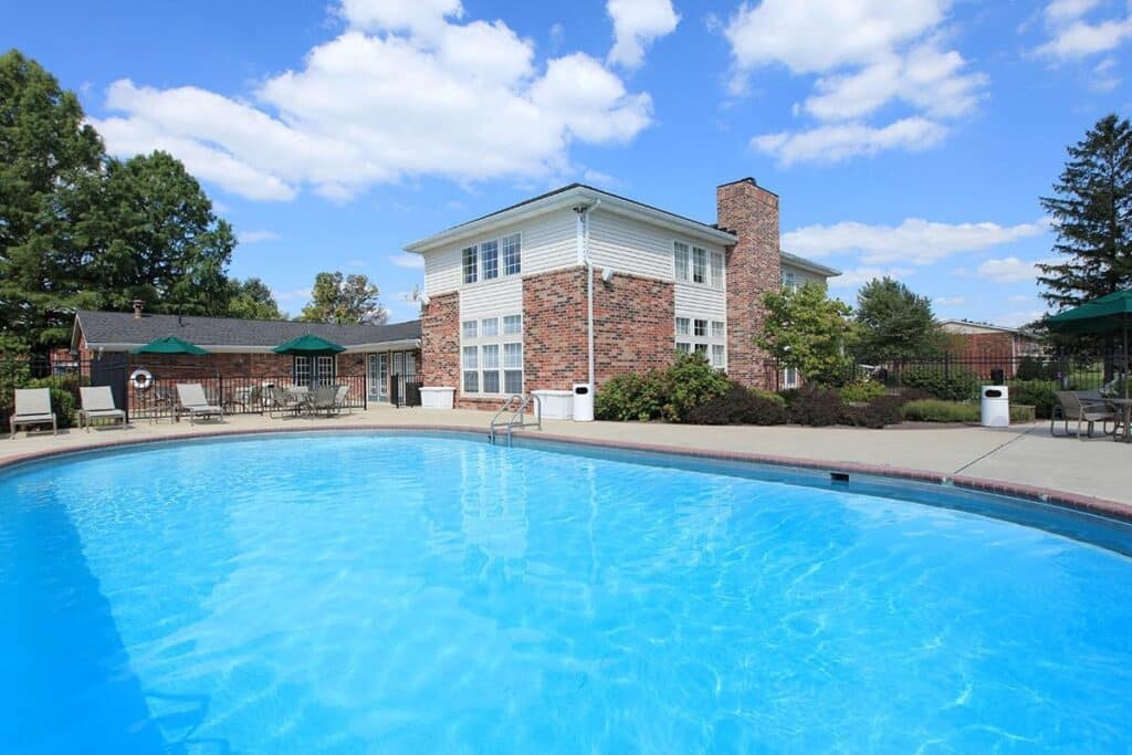 Outdoor swimming pool with clear blue water in front of a two-story brick building, surrounded by lounge chairs, tables, and green trees under a sunny sky.