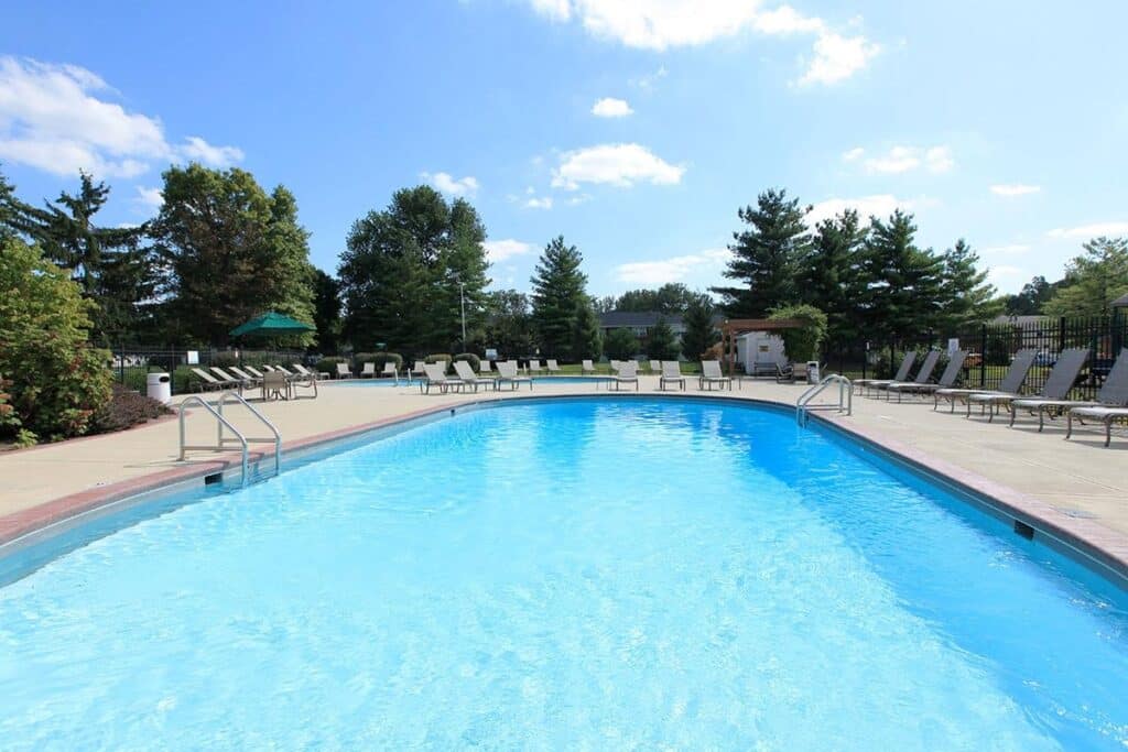 Outdoor swimming pool with clear blue water, surrounded by lounge chairs, trees, and a clear sky with scattered clouds.