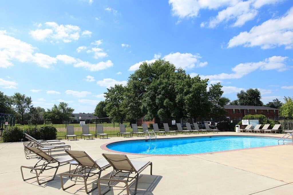 Outdoor swimming pool surrounded by lounge chairs, with trees and a fence in the background under a blue sky with scattered clouds.