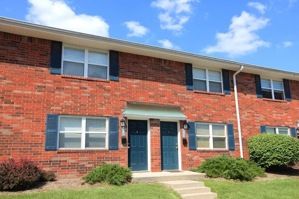 A two-story brick apartment building with blue shutters and two blue front doors, surrounded by shrubs and a grassy lawn under a blue sky.