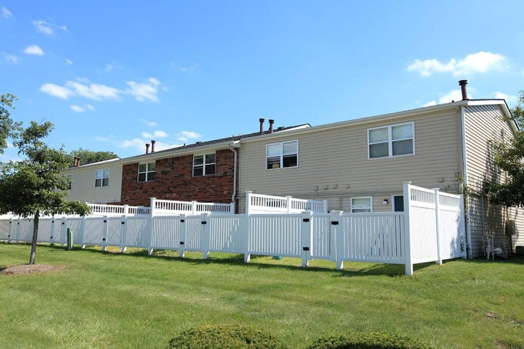 A row of two-story townhouses with beige and brick exteriors, white fences, and a grassy yard under a blue sky with scattered clouds.