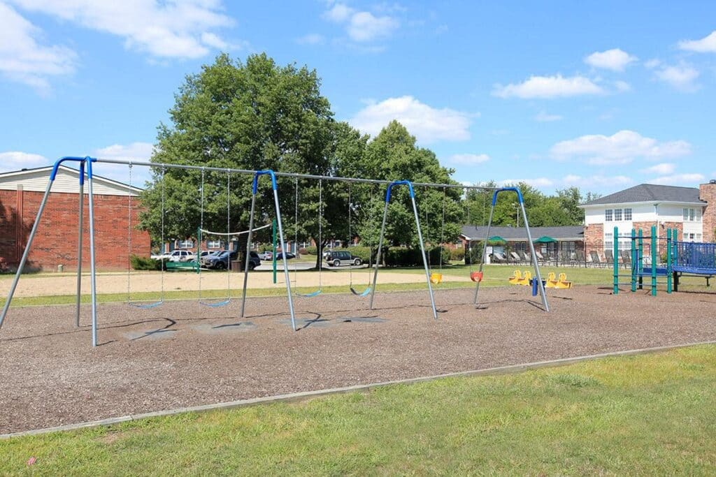A playground with empty swings, seesaws, and a slide, surrounded by grass and trees, with buildings and parked cars in the background.