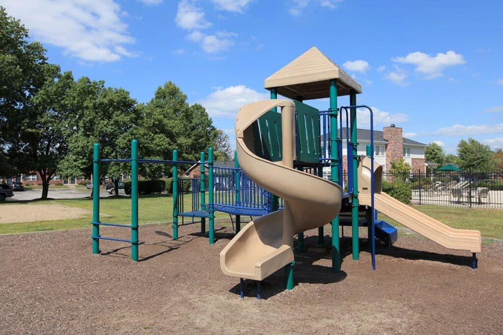 Outdoor playground with a spiral slide, climbing bars, and a small platform structure, situated on a mulch surface with grass and trees in the background.