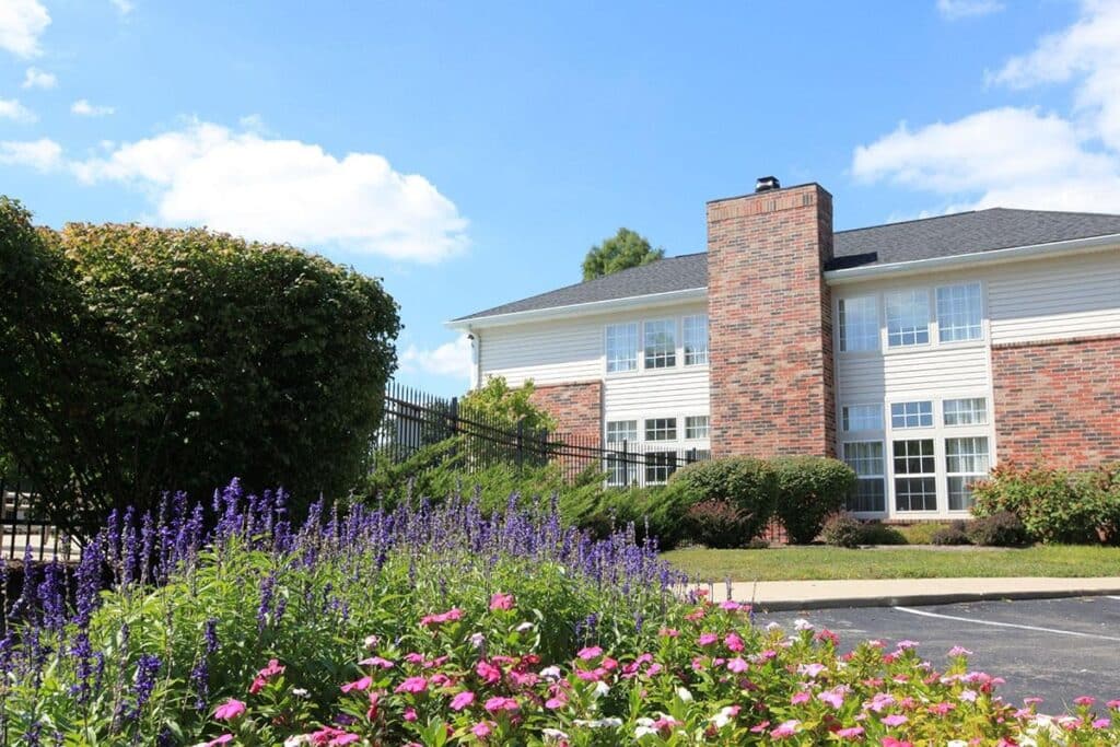 A brick building with large windows stands behind a garden filled with purple and pink flowers under a blue sky with scattered clouds.