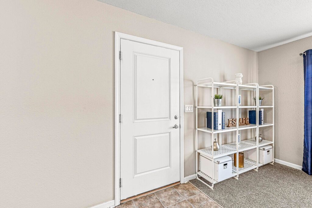 A white front door next to a white metal shelving unit holding books, plants, and decorative items in a carpeted entryway.