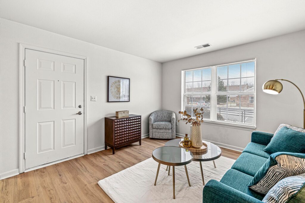 A living room with a teal sofa, two round glass coffee tables, a gray armchair, a wooden cabinet, a floor lamp, and a large window letting in natural light.