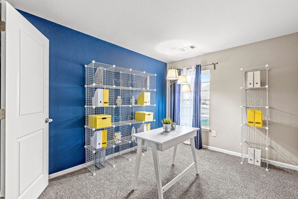 A modern home office with a white desk, metal wire shelving units holding yellow and white storage boxes, blue accent wall, floor lamp, and window with blue curtains.