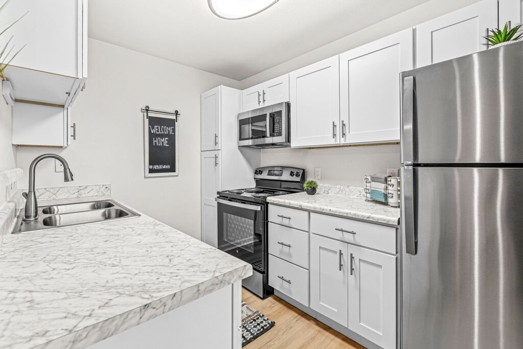 Modern kitchen with white cabinets, marble countertops, stainless steel appliances, double sink, and a “Welcome Home” sign on the wall.