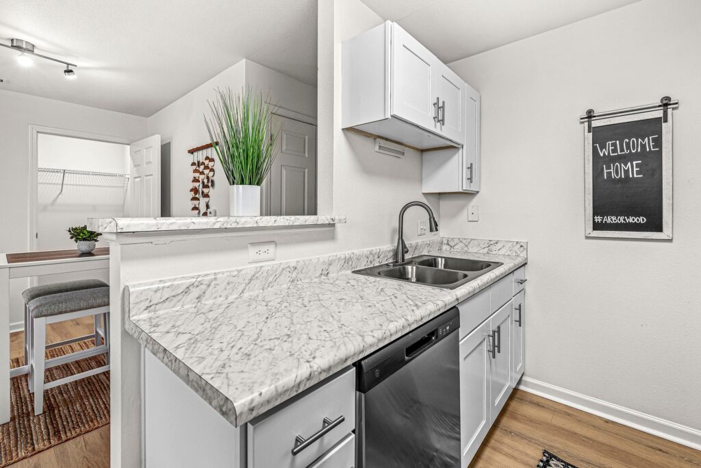 Modern kitchen with marble countertops, stainless steel sink and dishwasher, white cabinets, and a "Welcome Home" sign on the wall. A plant decorates the counter.