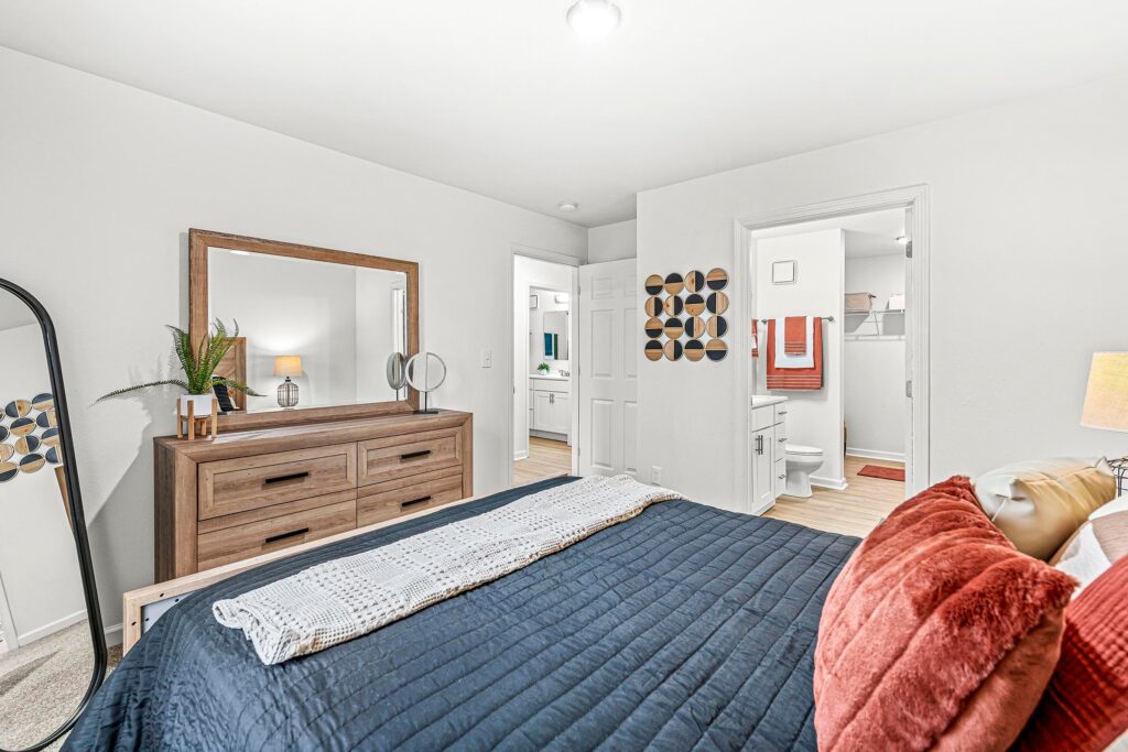 Bedroom with a bed, dresser, large mirror, and view into an adjoining bathroom; decor includes wall art and houseplants.