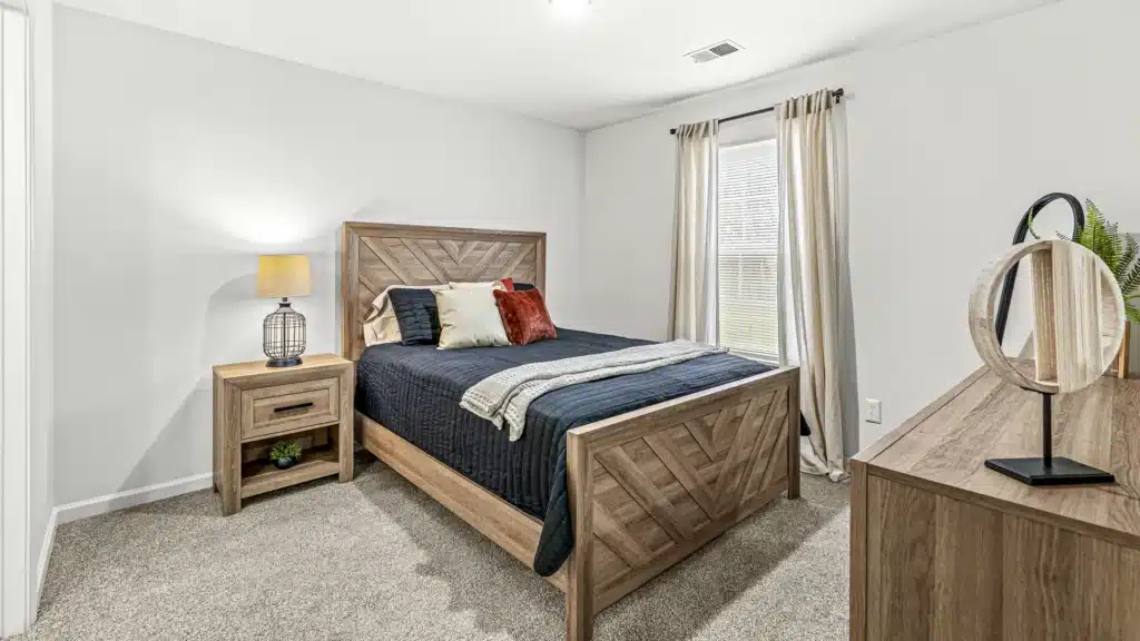 A bedroom with a wooden bed frame, black bedding, two pillows, a nightstand with a lamp, a dresser with decor, beige carpet, and a window with light curtains.