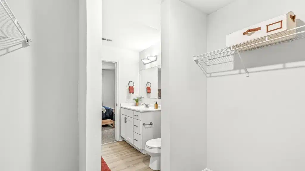 White bathroom with a vanity, toilet, mirror, wall shelves, and a glimpse into an adjacent bedroom. The space features light wood flooring and minimal decor.