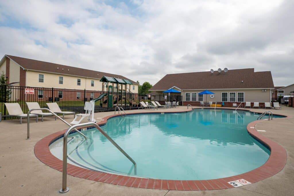 Outdoor swimming pool with metal handrails, lounge chairs, and a playground in the background, adjacent to a residential building and clubhouse under a cloudy sky.